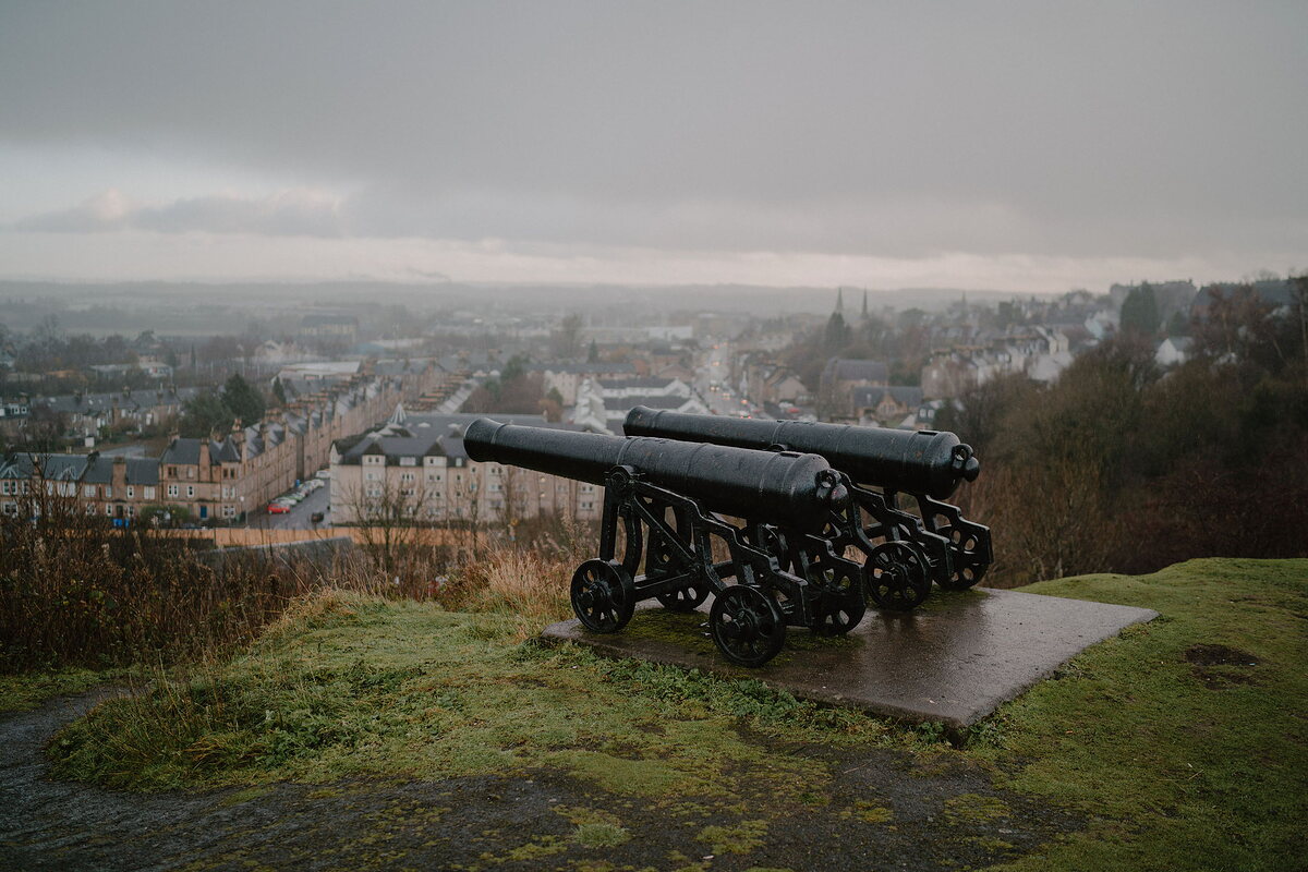Two black cast-iron cannons point out from a grassy hilltop over a sprawling town below, mist obscuring the distant hills.