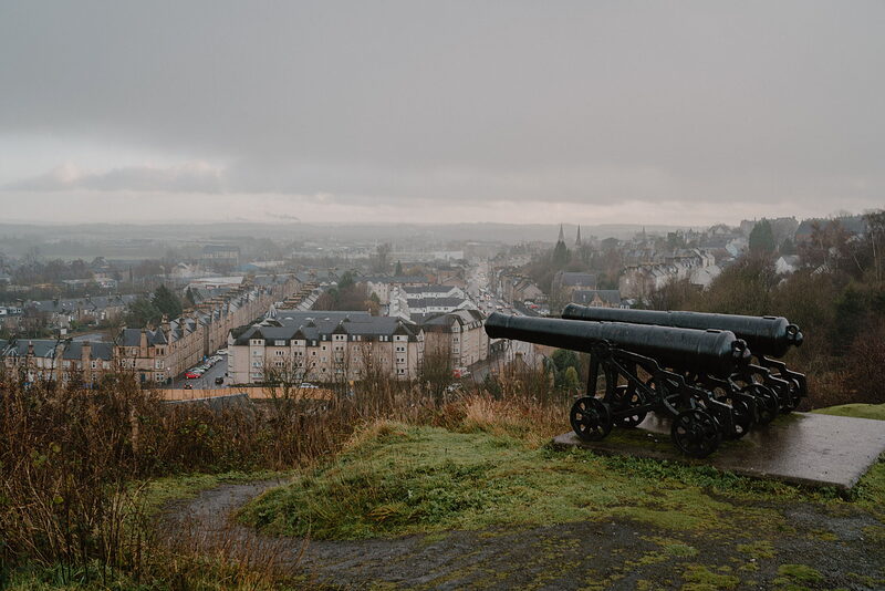 Two black cast-iron cannons point out from a misty hilltop over a sprawling town of terraced streets below.