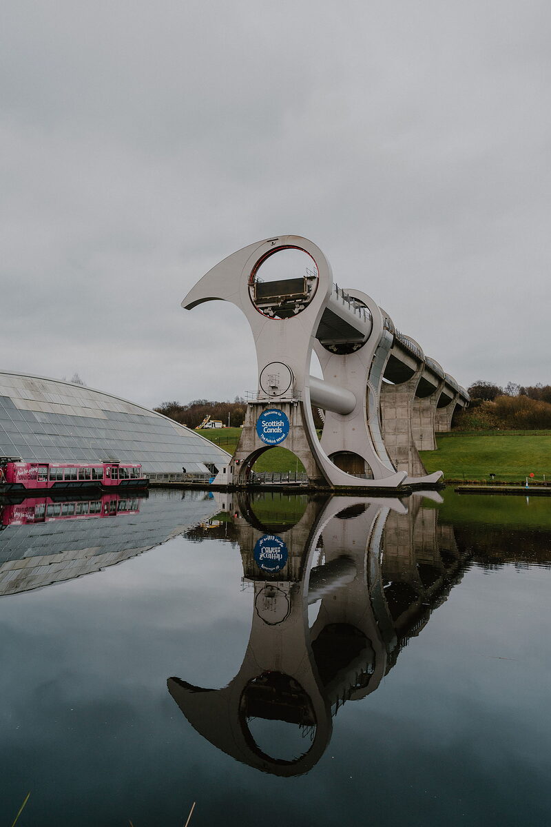 The curved white superstructure of a rotating boat lift rises beside a glass-domed visitor centre, both reflected in the still canal water, a pink narrowboat moored alongside.