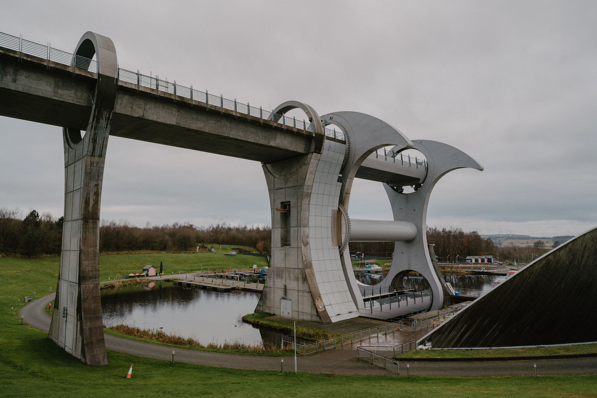 The curved white superstructure of a rotating boat lift arches over a canal basin and a stone aqueduct, a grass lawn in the foreground.