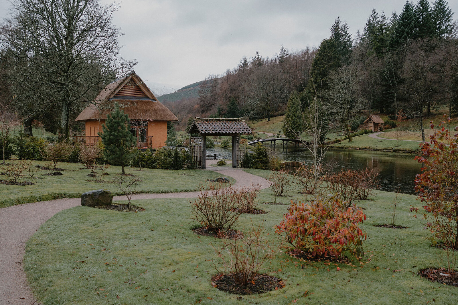 A gravel path winds past newly planted shrubs toward a thatched cabin and a timber gateway beside a still pond.