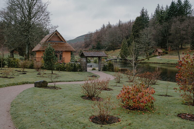 A gravel path winds past newly planted shrubs toward a thatched cabin and a timber gateway beside a still pond.