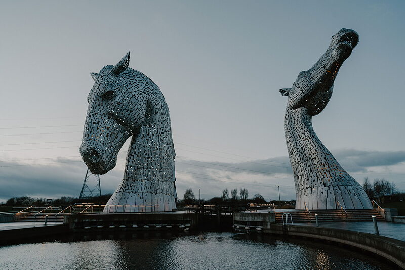 One of the towering steel horse-head sculptures known as the Kelpies rears beside its twin against a dusk sky, reflected in the canal water.