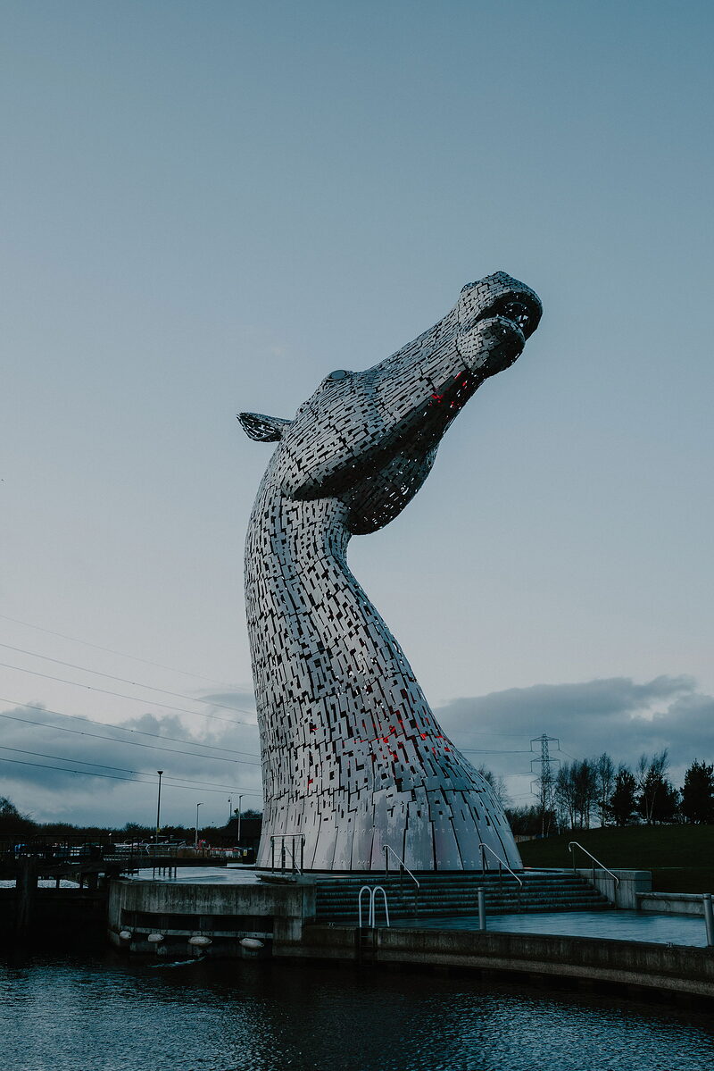 One of the towering steel horse-head sculptures known as the Kelpies rears against a dusk sky, steps leading down to the water below.