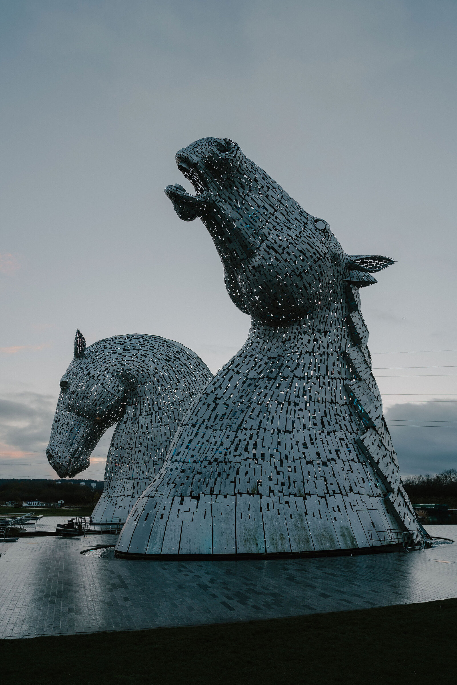 The towering steel horse-head sculptures known as the Kelpies rear together against a dusk sky, water lapping at their base.