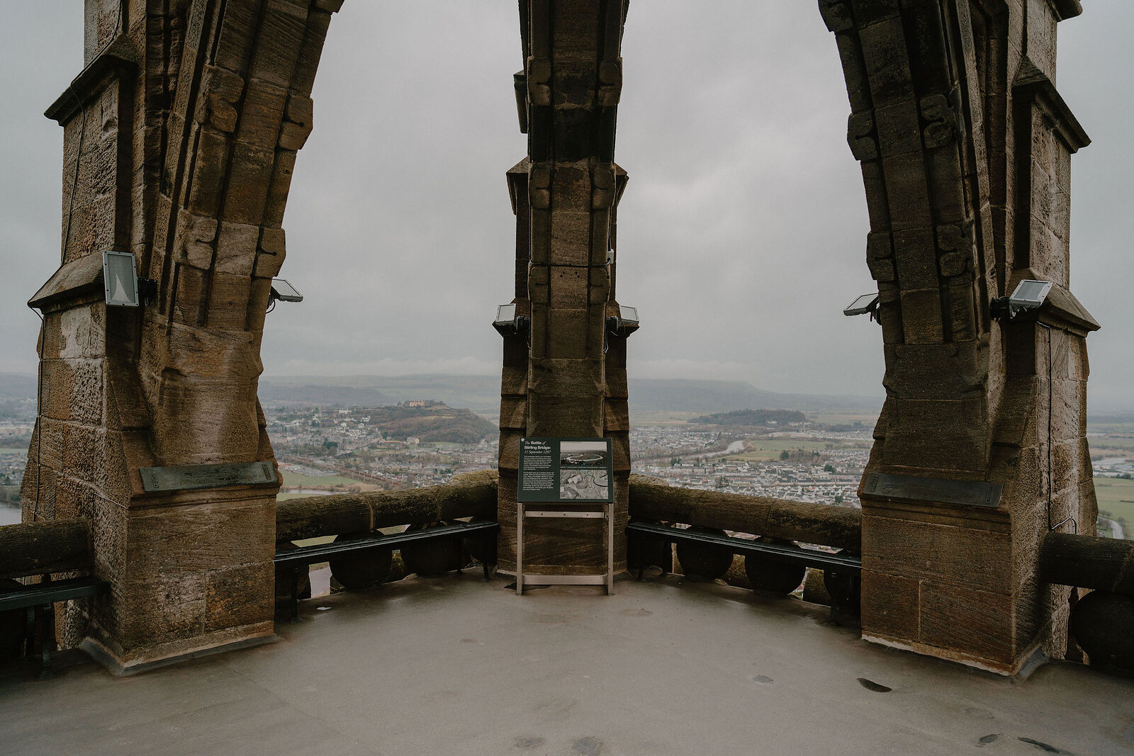 Three tall stone arches frame a panoramic view of a town and hills from an elevated monument gallery, benches lining the walls beneath.