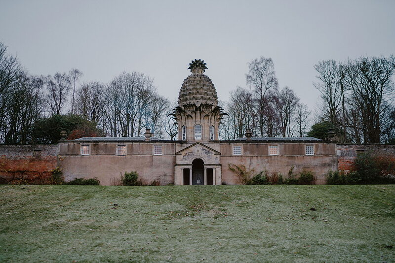 A distinctive stone folly shaped like a giant pineapple crowns a walled garden building, frosted grass stretching in front.