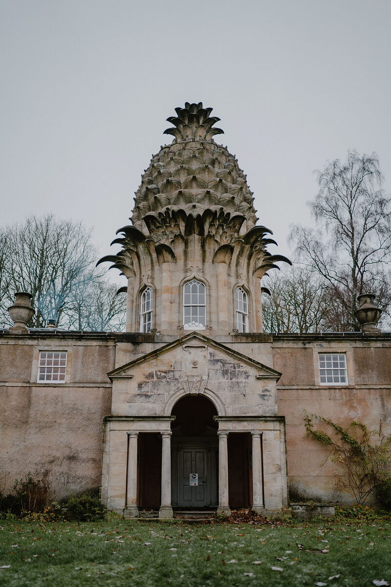A distinctive stone folly shaped like a giant pineapple rises against a pale sky, arched windows set into its base.