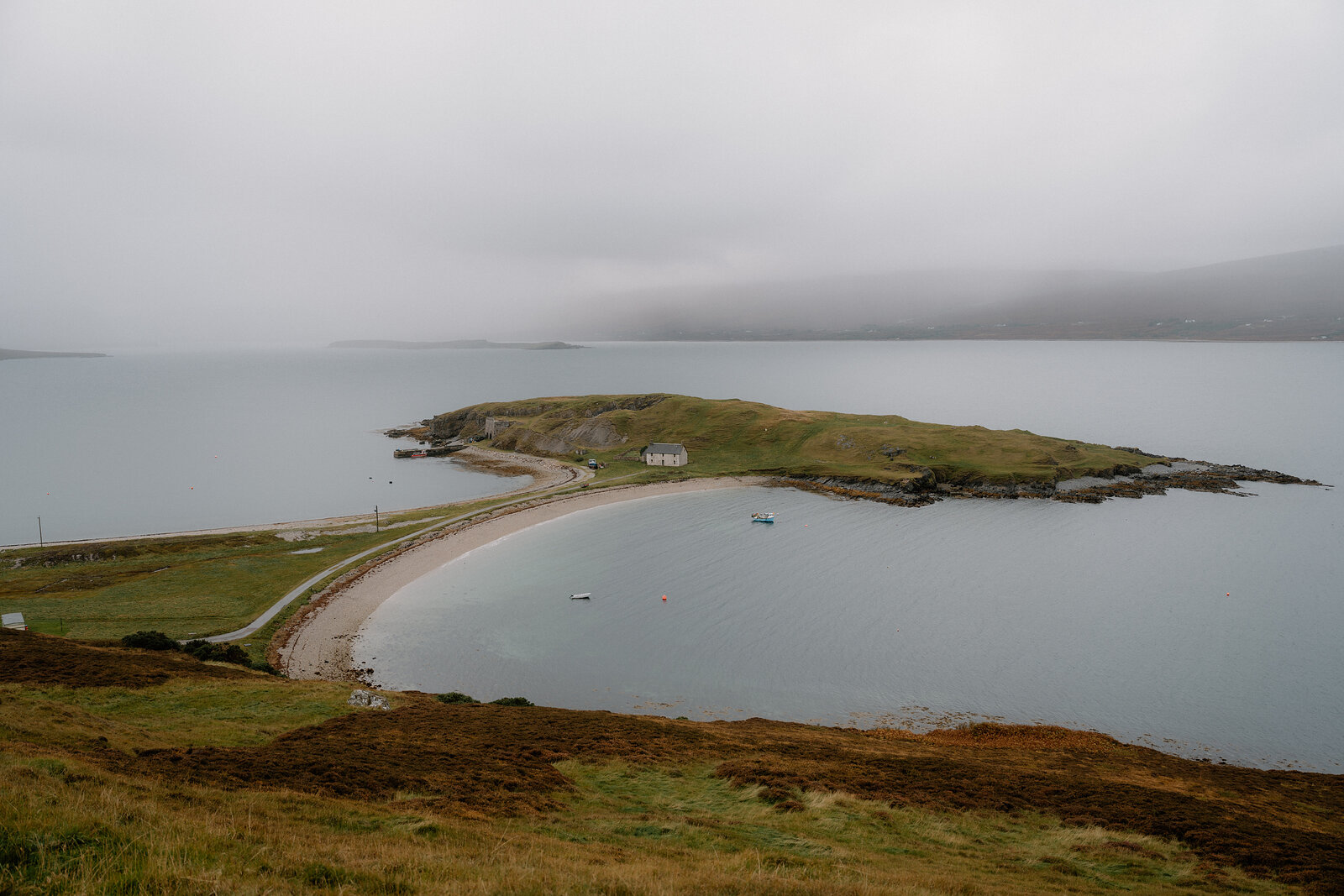 A curving sandy spit connecting a grassy headland to the shore, mist rolling over the hills beyond