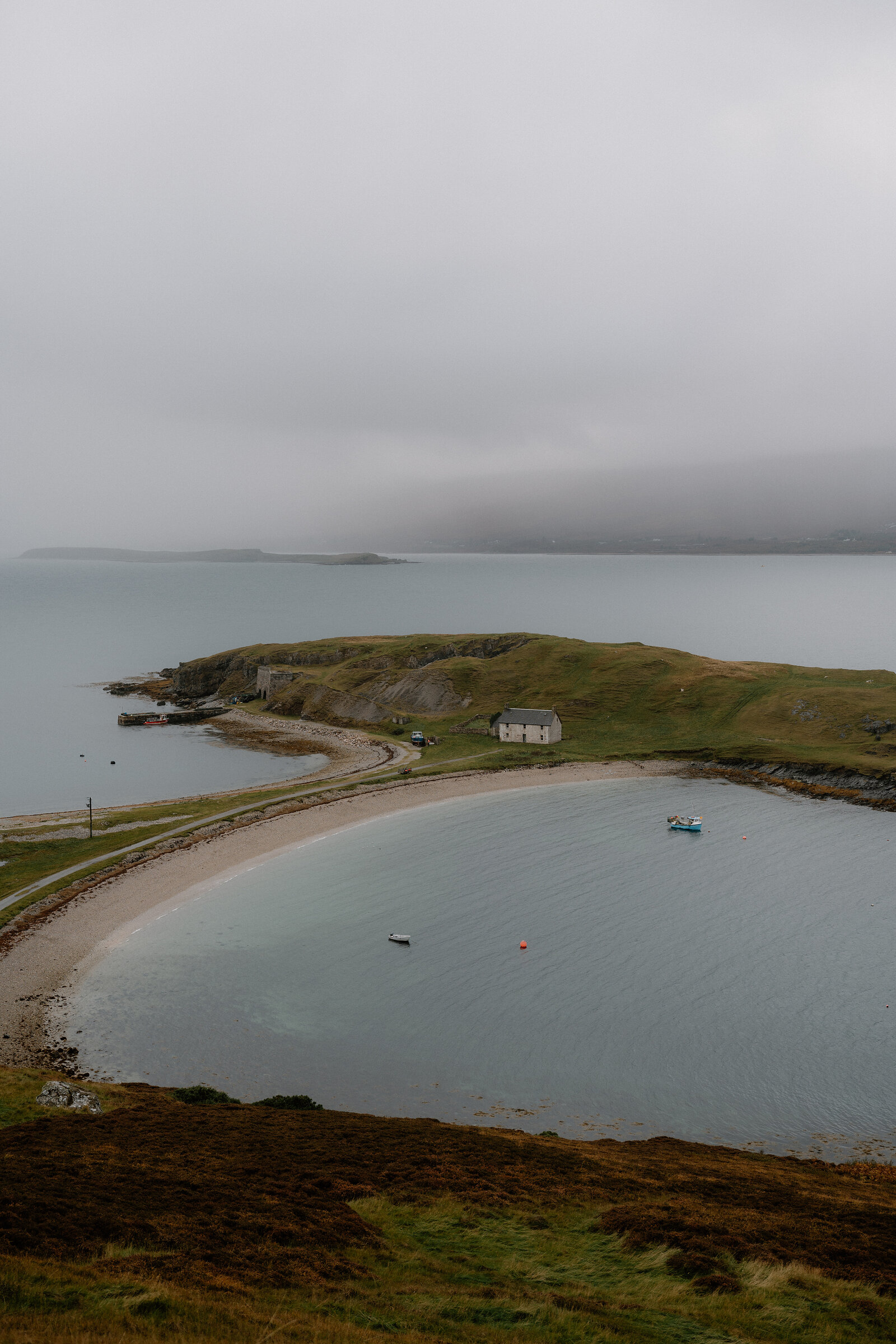 A curving sandy beach enclosed by a grassy headland, small boats anchored in the calm water