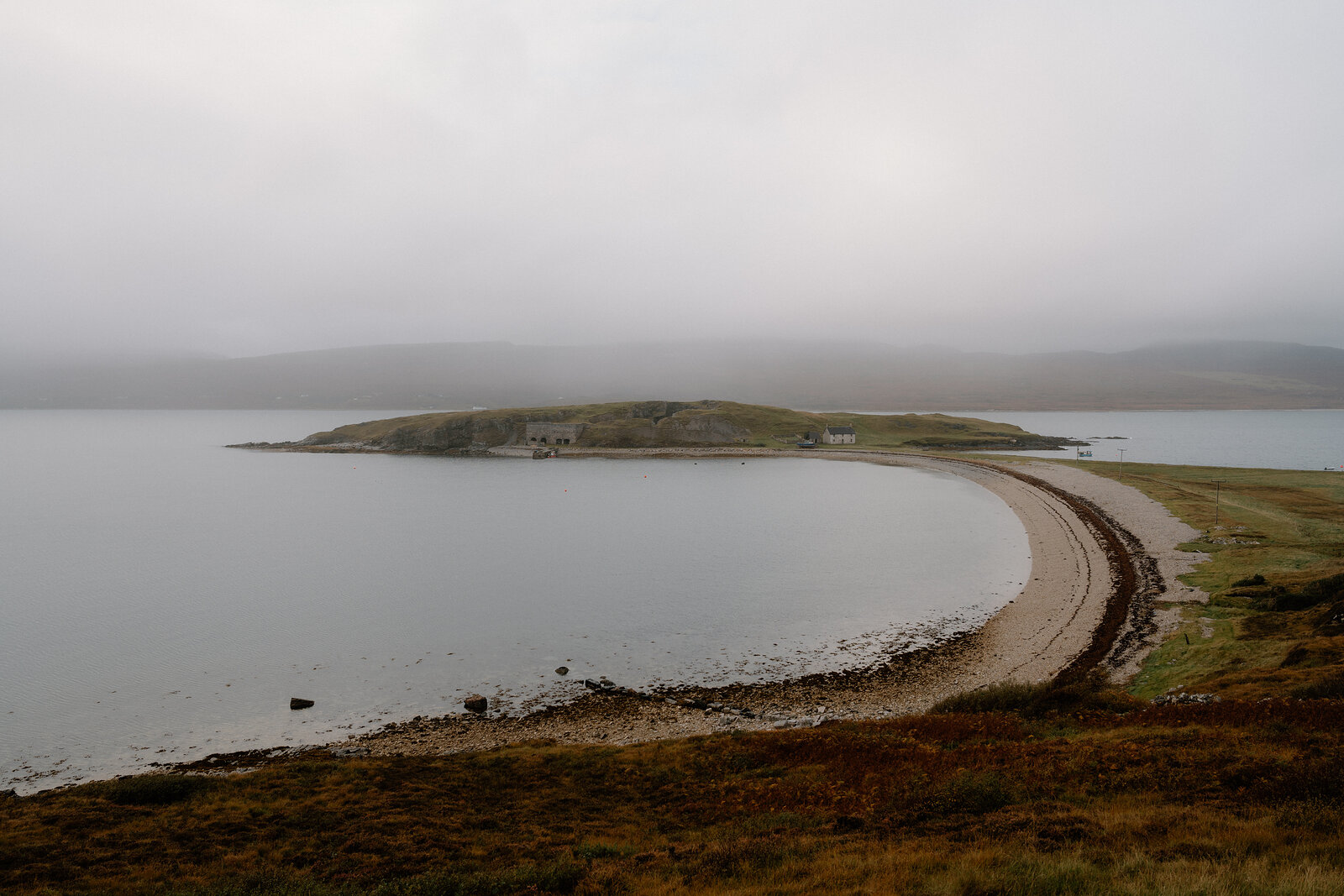 A curving sandy spit connecting a grassy headland to the shore, mist rolling over the hills beyond