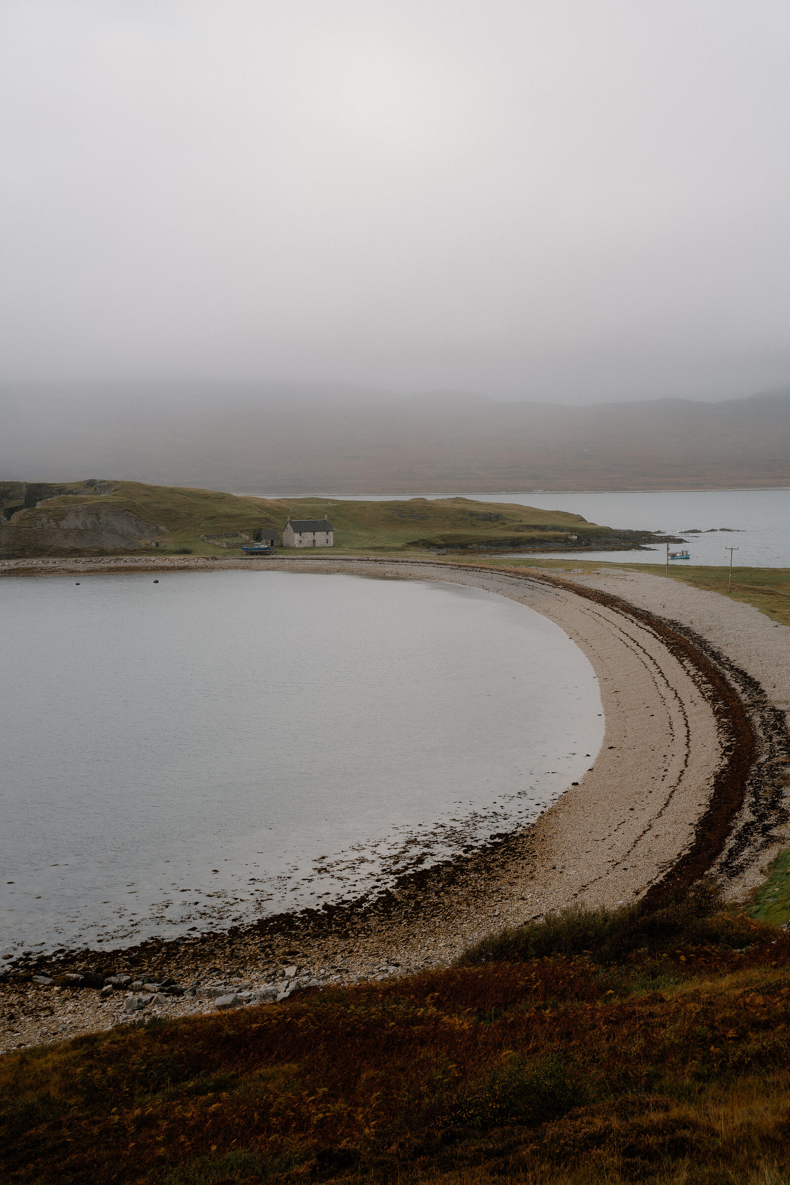 A curving shingle beach enclosed by a grassy headland, a house set on the shore beneath mist-covered hills