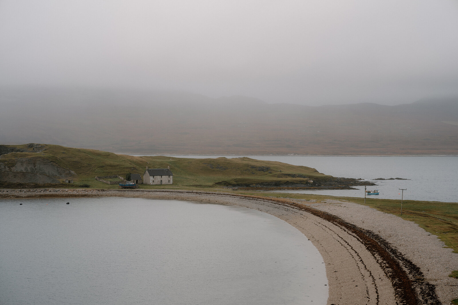 A curving sandy beach enclosed by a grassy headland, a house set on the shore beneath mist-covered hills
