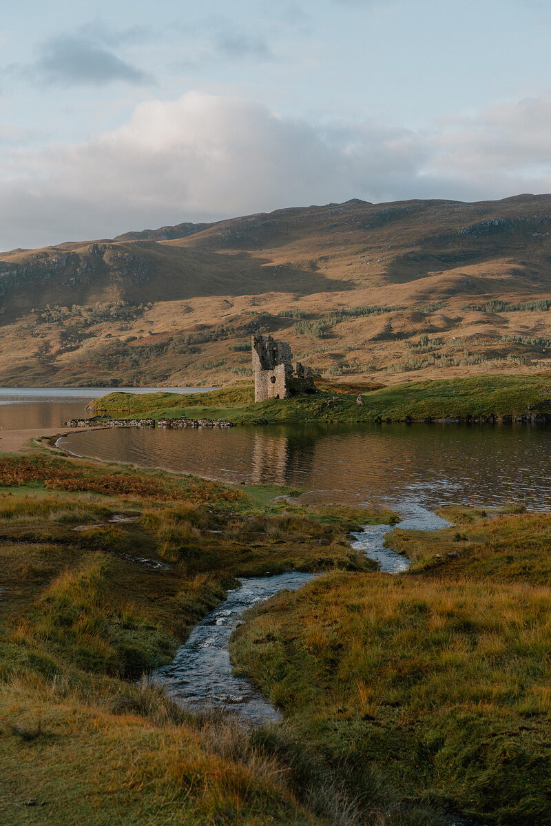 A ruined castle standing on the shore of a loch, a stream winding through the grass before it.