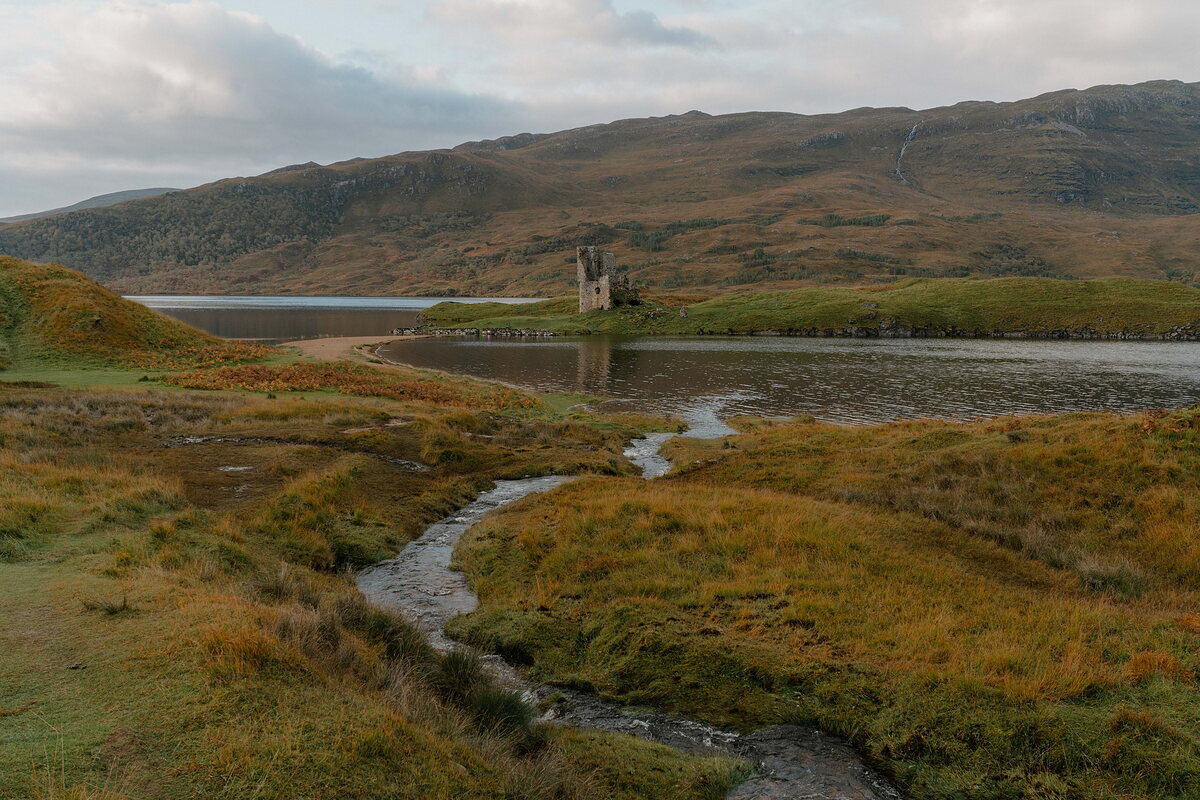 A ruined castle standing on the shore of a loch, a stream winding through the grass before it.
