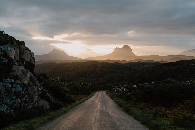 A single-track road winding towards distinctive mountain peaks silhouetted against a glowing sunset.