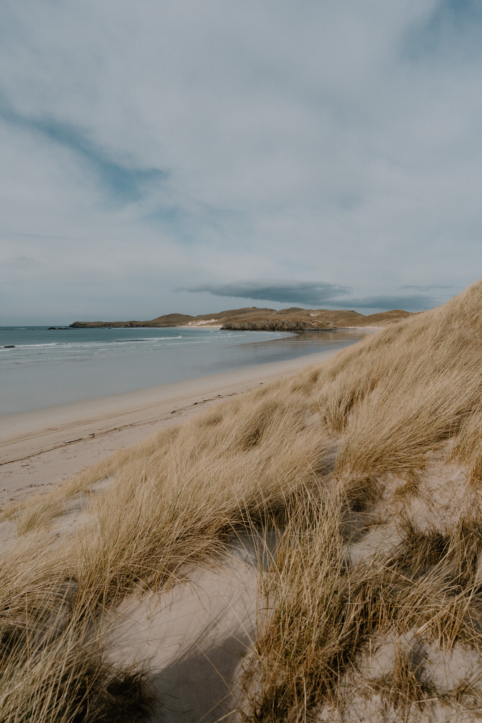 A sandy beach curving beneath dune grass, small islands visible offshore