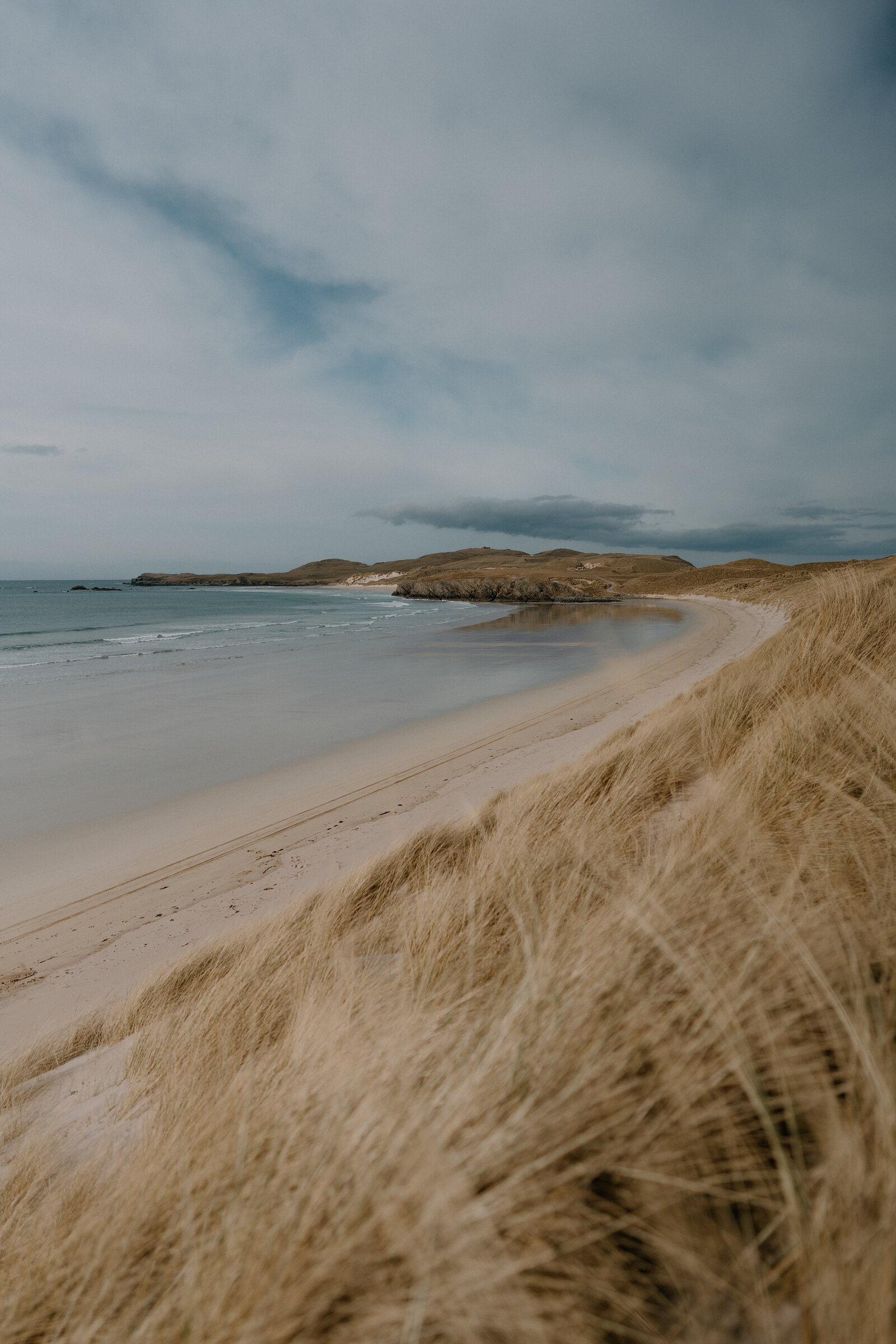 A curving sandy beach backed by dune grass, hills rising at the far end of the bay
