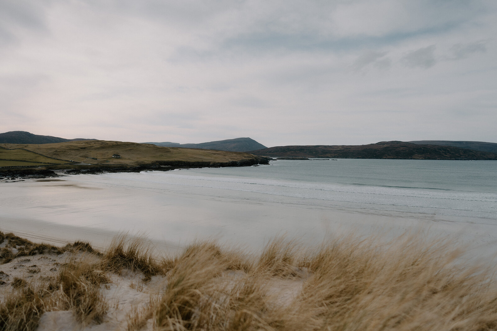 A curving sandy beach backed by dune grass, hills fading into the distance across the bay