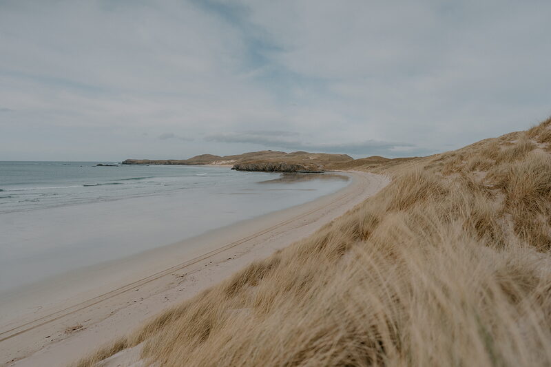 A curving sandy beach backed by dune grass, a rocky headland closing the bay.