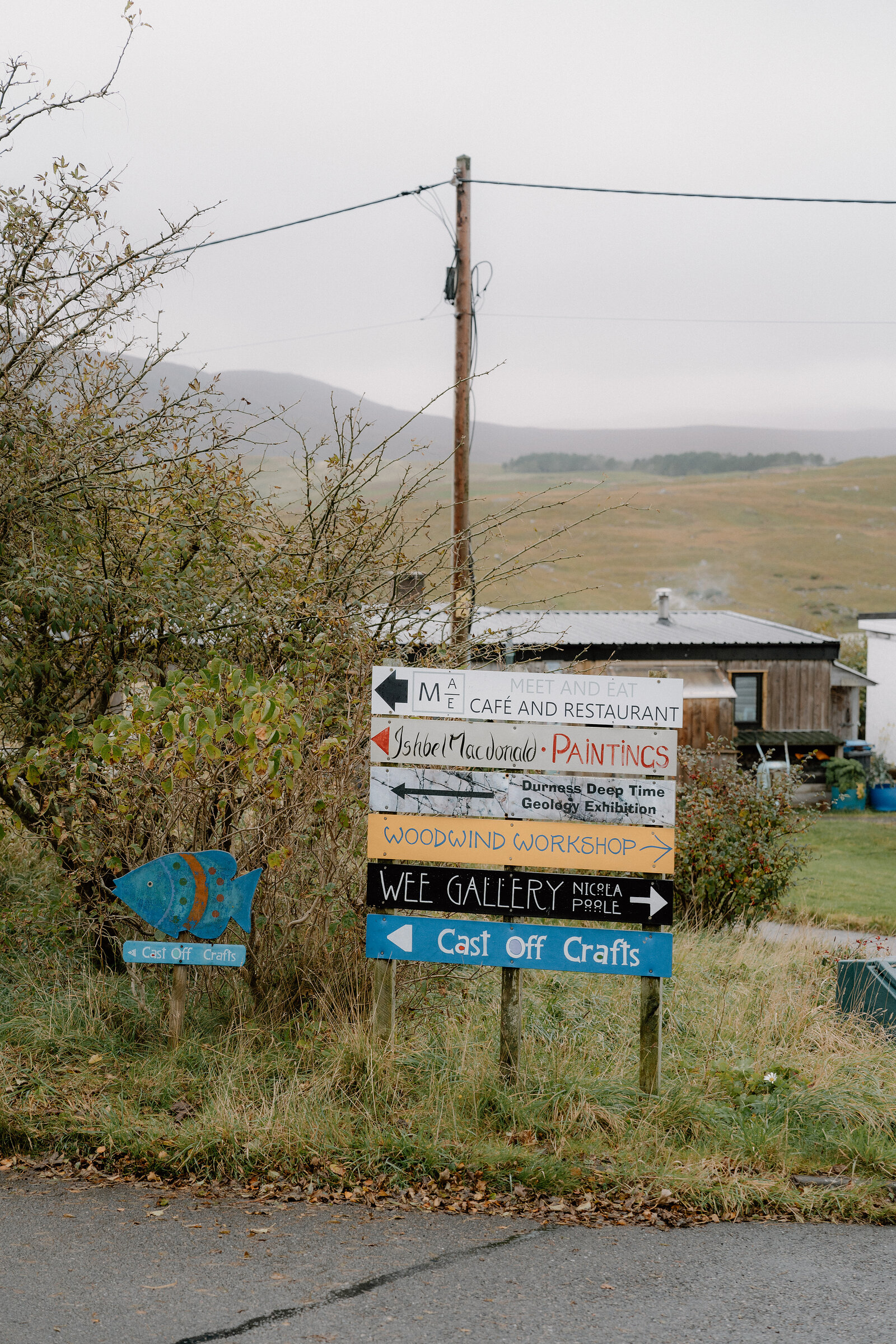 A cluster of hand-painted signs pointing to a cafe, gallery and craft workshops beside a country road