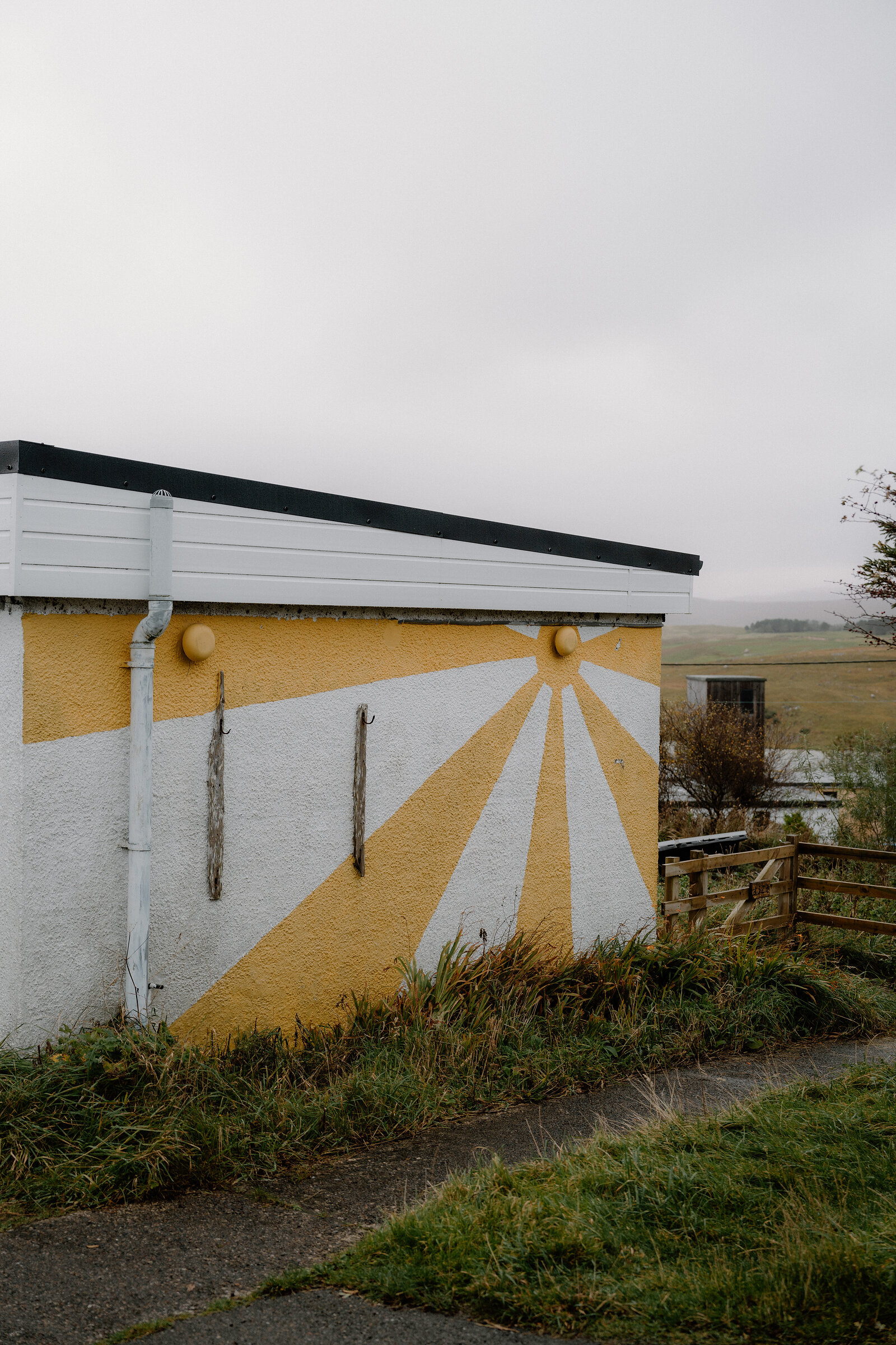 A sun mural painted on the gable end of a white building, fields stretching beyond