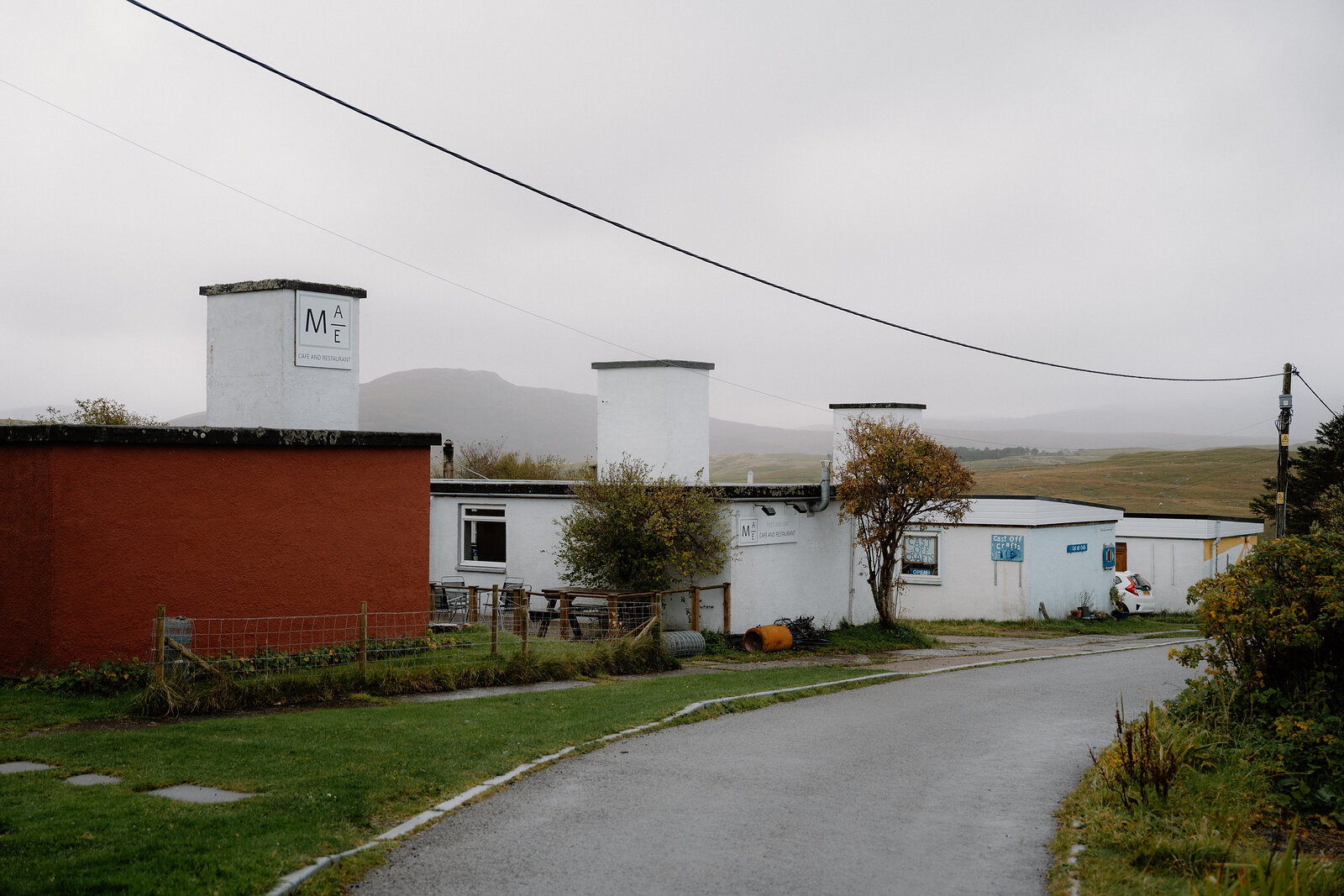 A row of chimney-topped white buildings along a country road, hills rising behind under mist