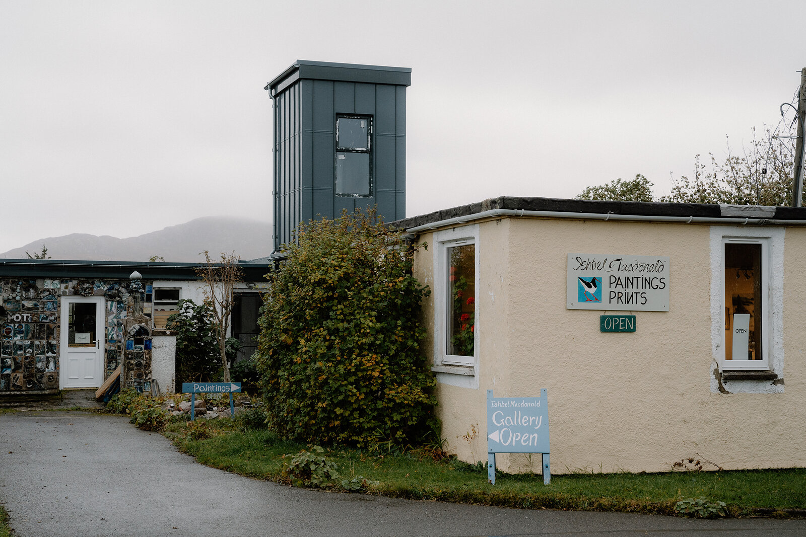 A gallery building with signs reading Paintings, Prints and Gallery Open beside a tall dark tower