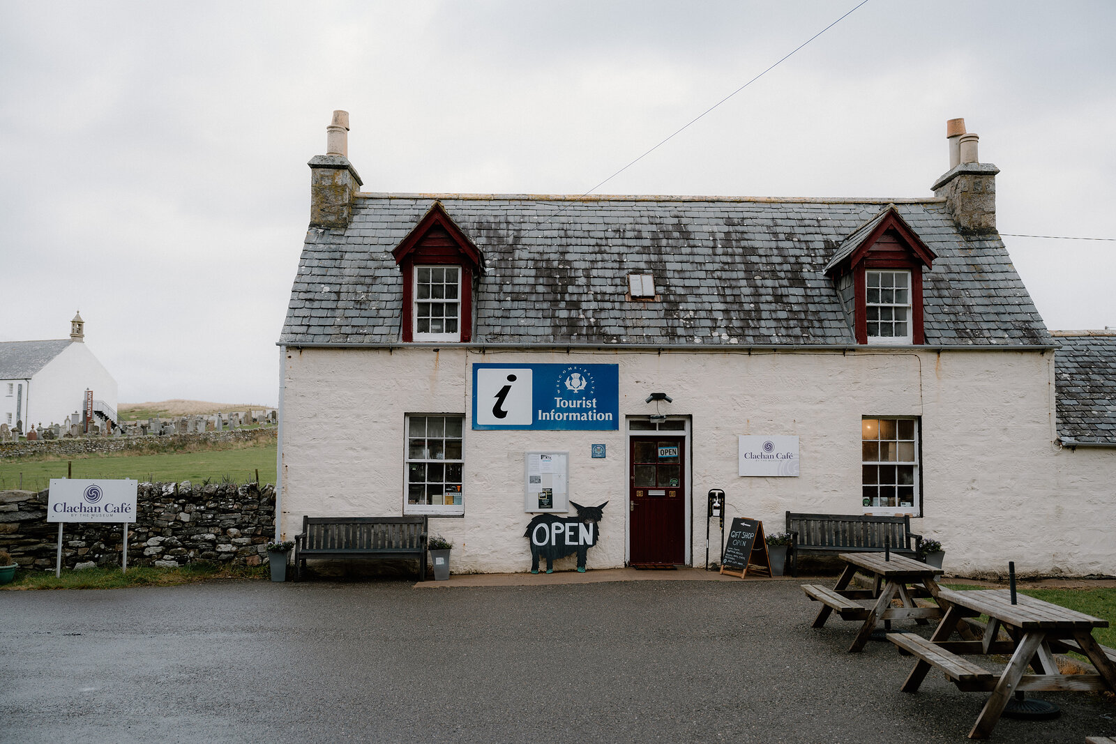 A white cottage with a Tourist Information sign and an Open board, picnic tables set outside