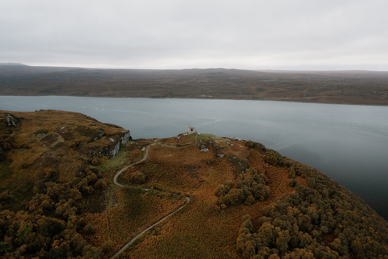 An aerial view of a wooded headland reaching into a calm loch, a small tower on its crest.