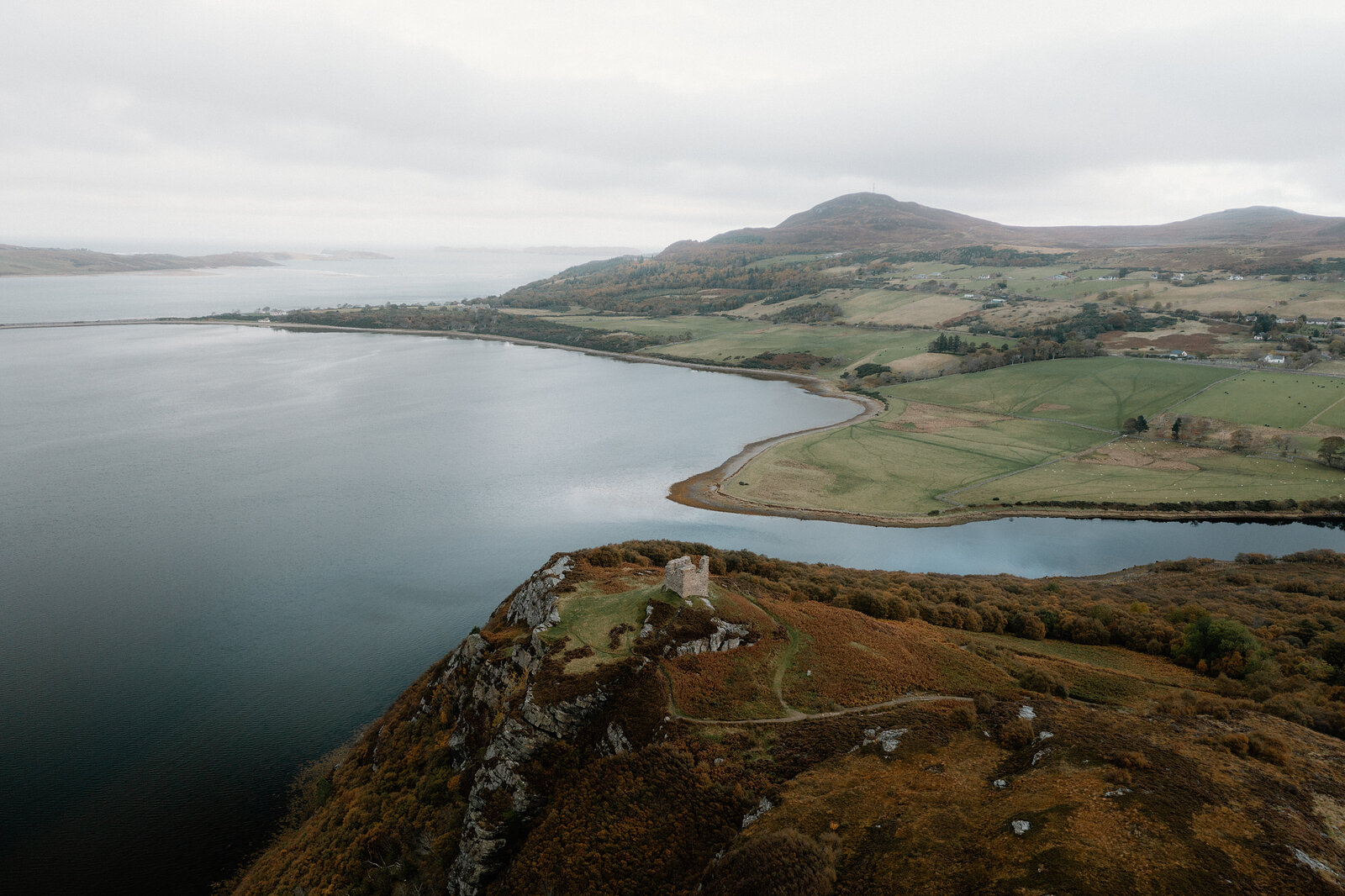 An aerial view of a loch shoreline curving past fields and a wooded headland, hills rising beyond