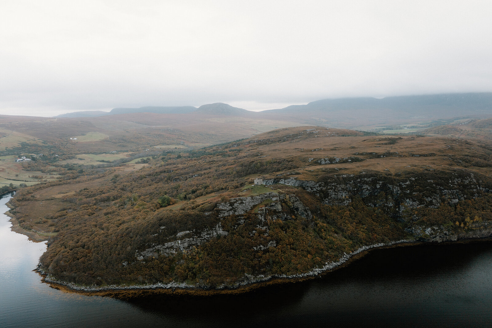 An aerial view of a wooded headland reaching into a loch, hills fading into mist behind