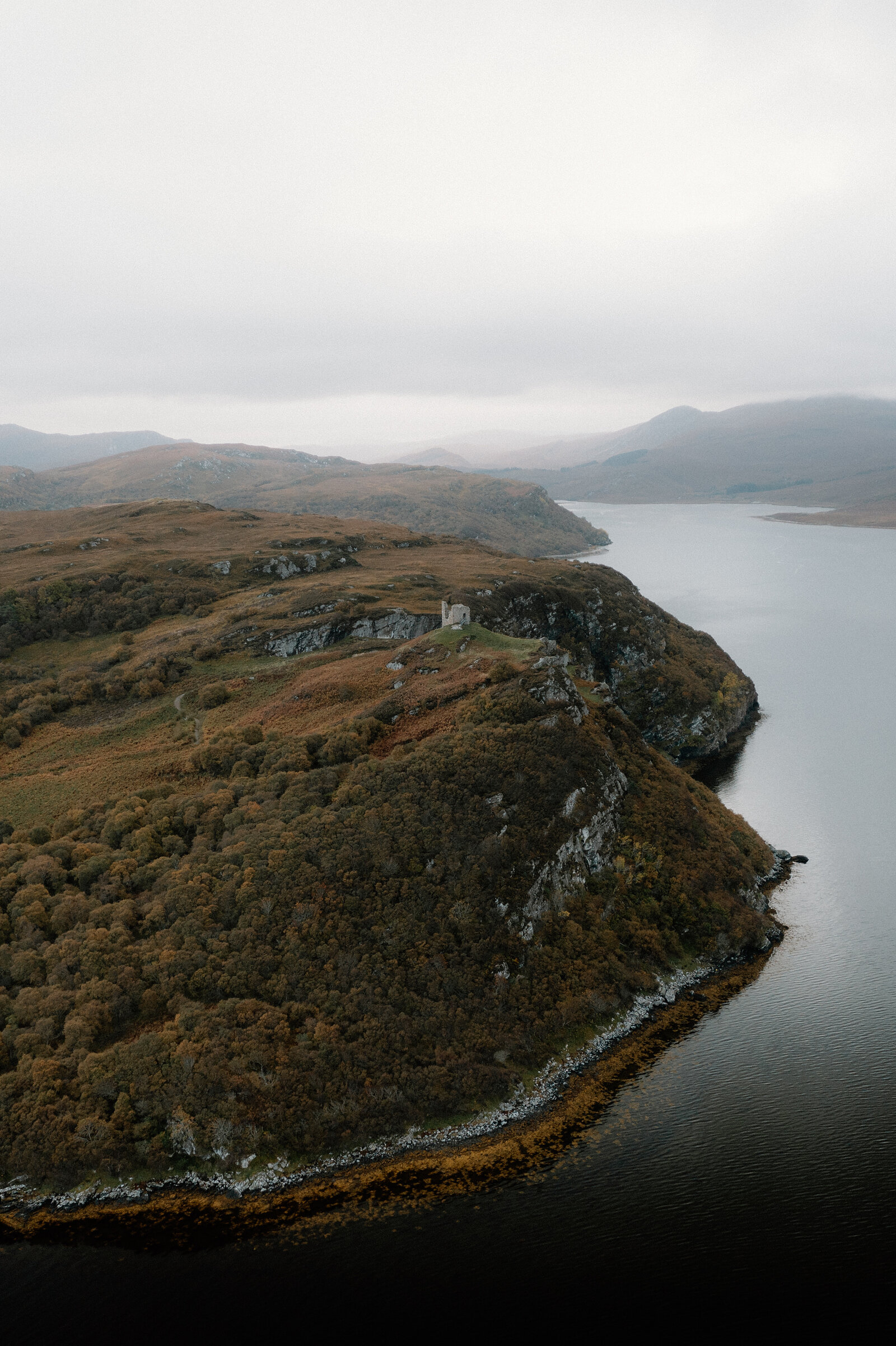 A wooded headland with a small ruin on its crest, reaching into a calm loch under low cloud
