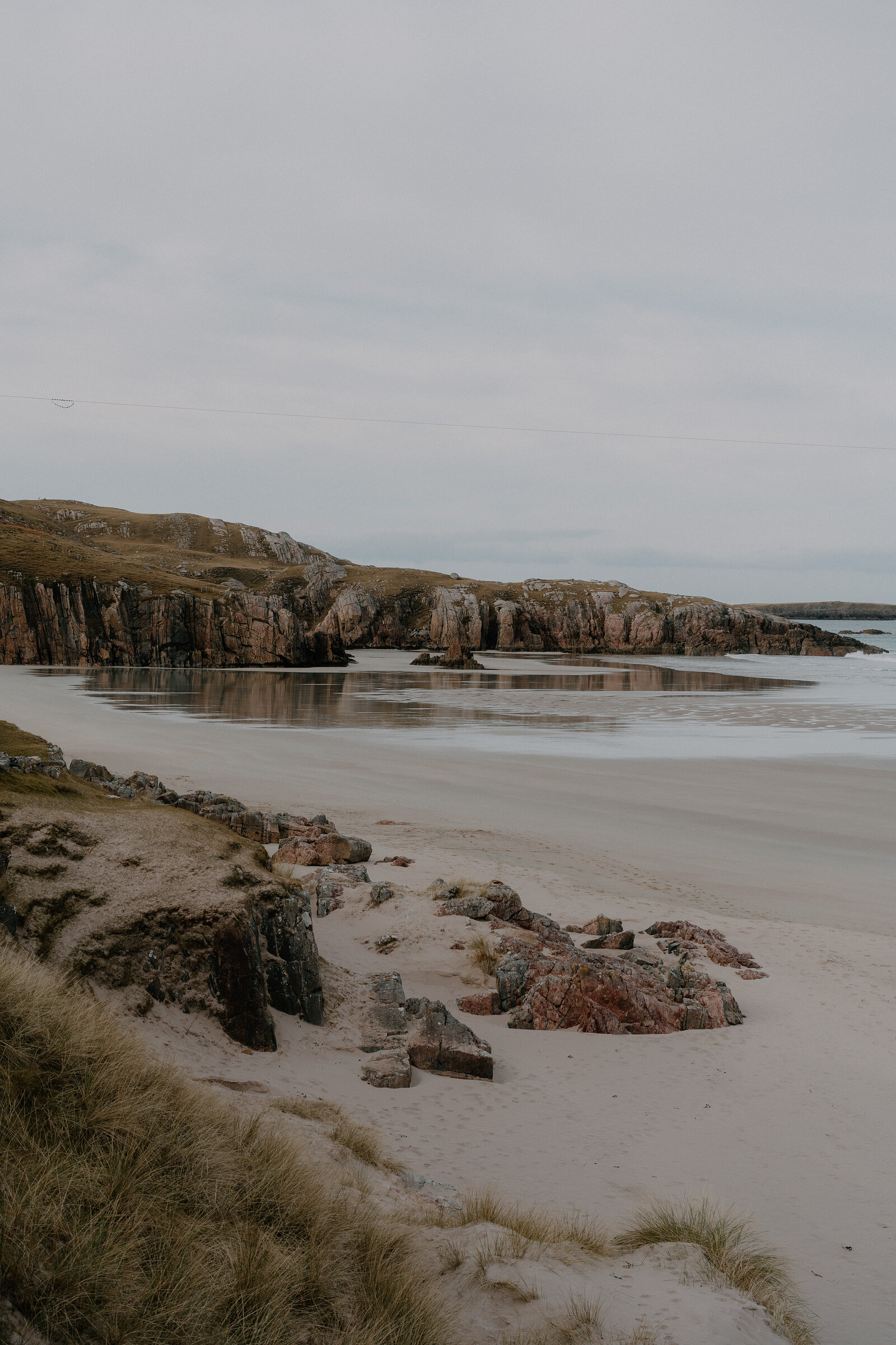 A sandy beach edged with pink-streaked rocks, cliffs reflected in the calm water