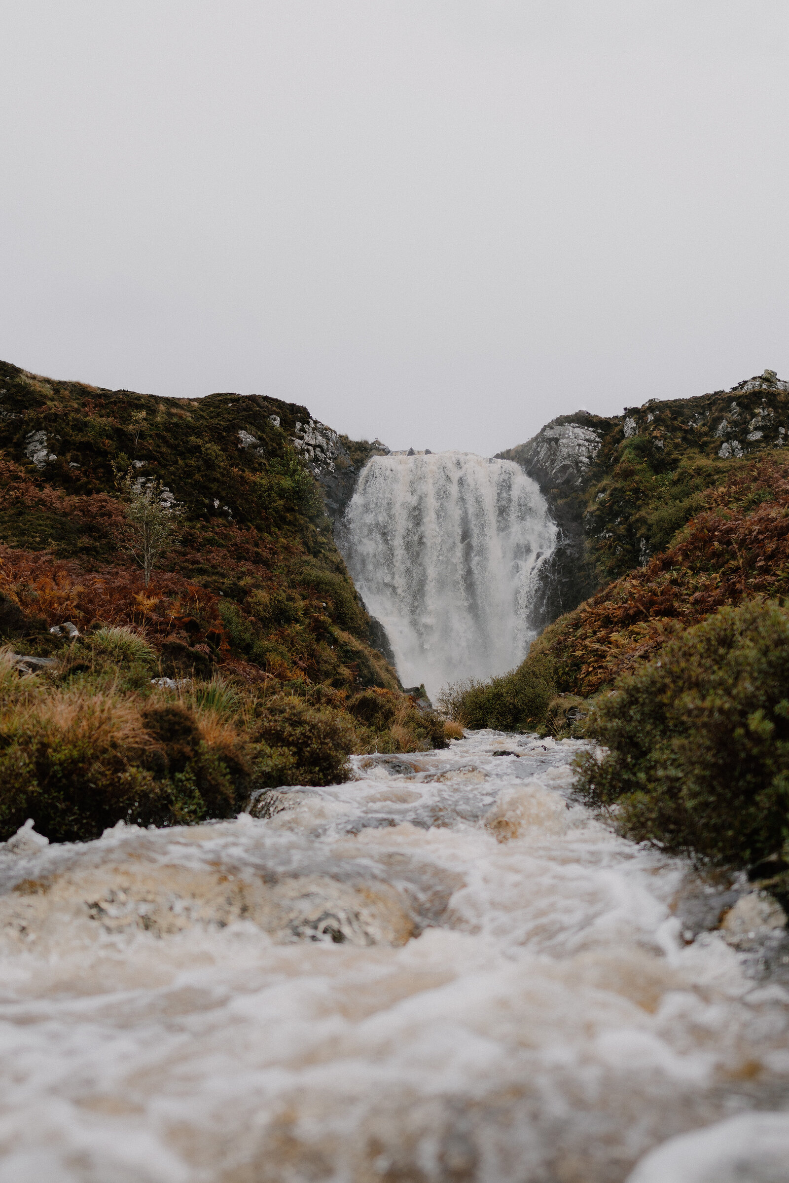 A waterfall dropping between rocky banks into a fast-flowing burn, autumn colour on the slopes