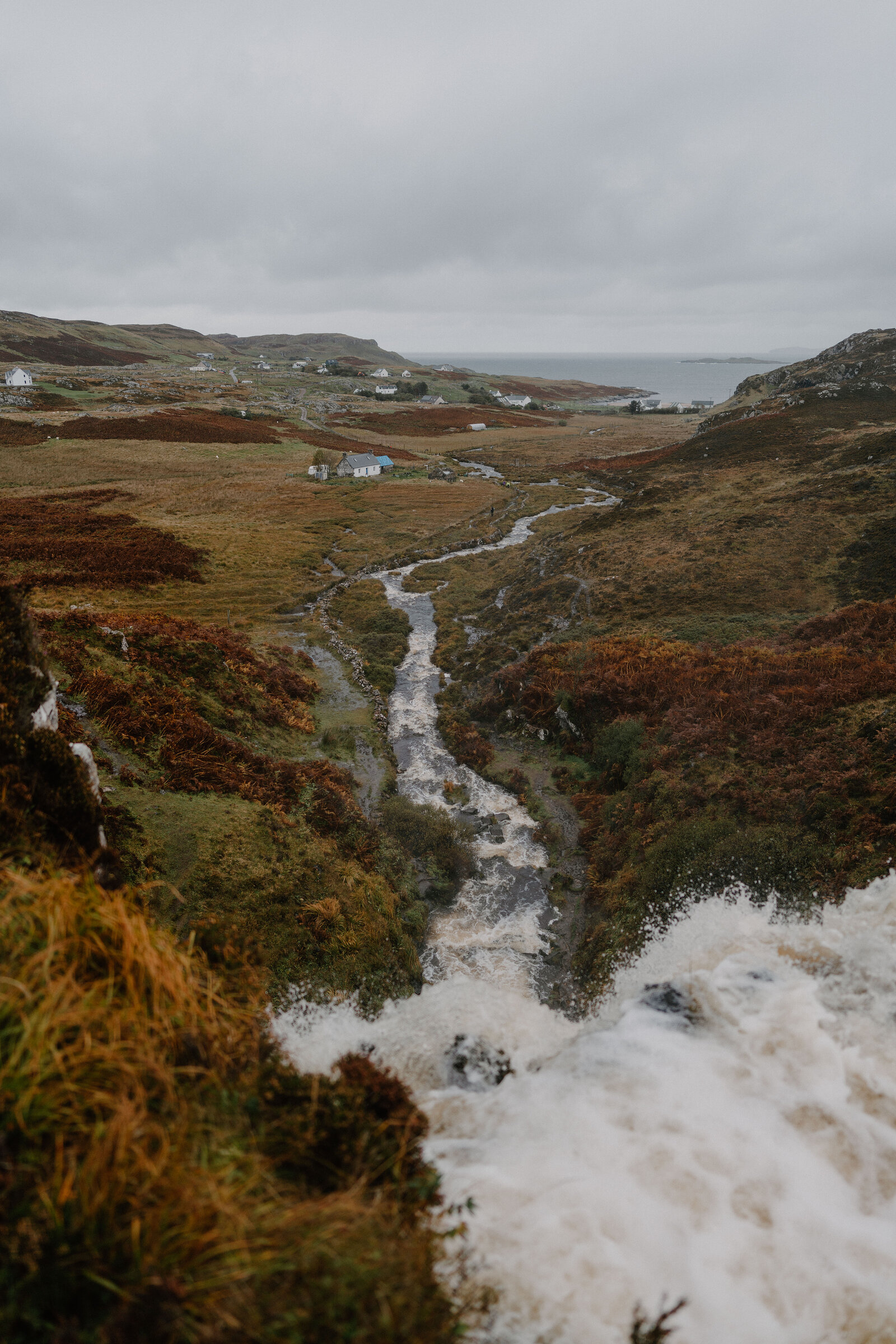 A fast-flowing burn cutting through heather-covered moorland towards a distant village and sea