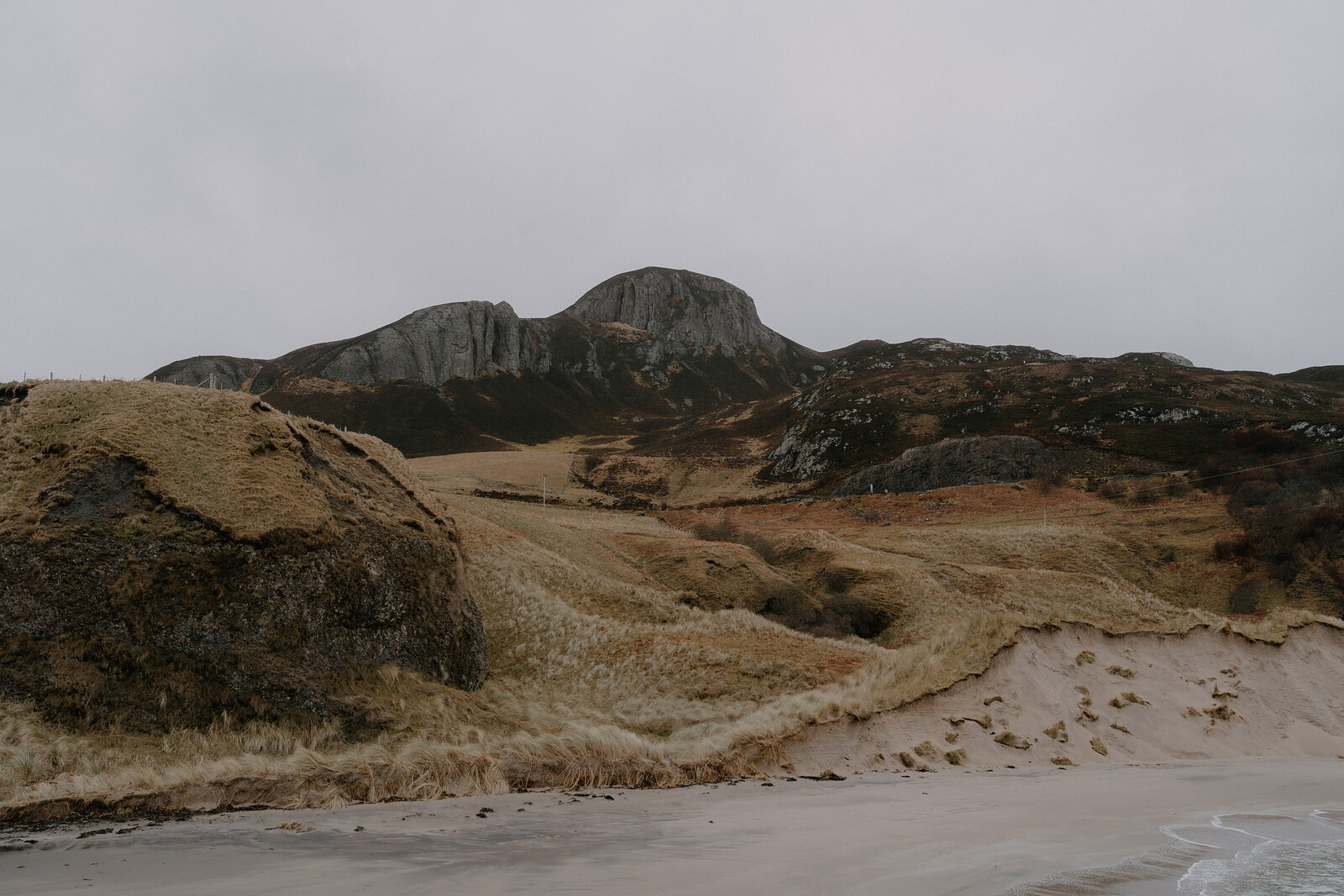 Grassy dunes lead to a rocky peak above a stretch of sandy beach.