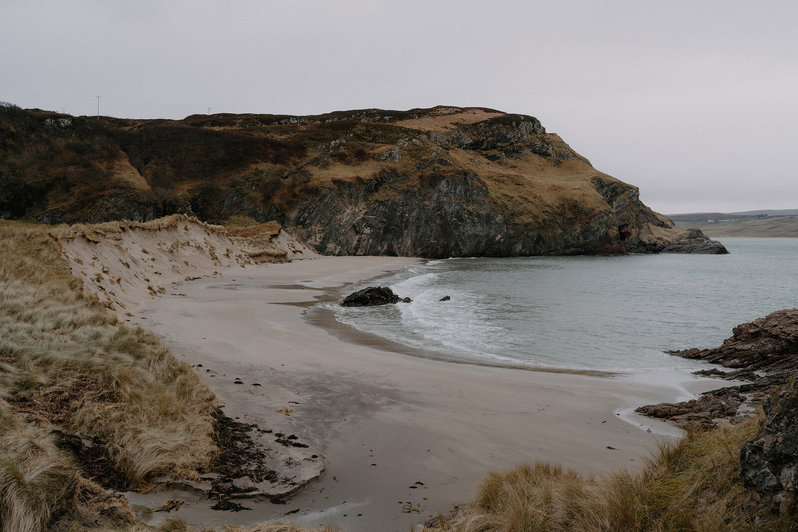 A crescent of sandy beach curves beneath rocky cliffs and grassy dunes.