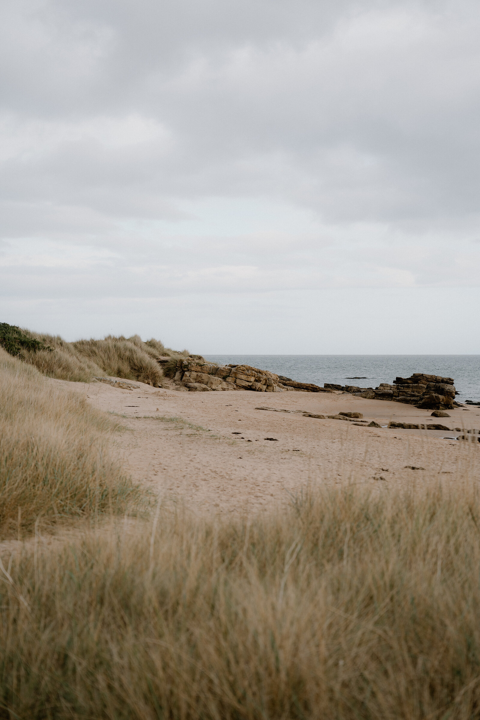 Rocky outcrops rise from a sandy beach fringed by dune grass.