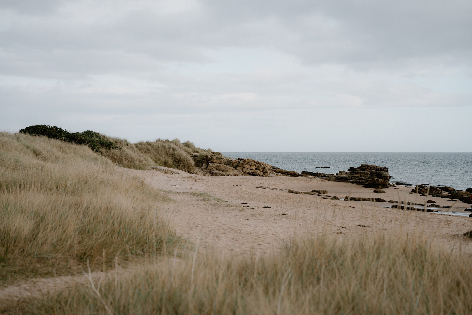 A sandy path through dune grass leads to a rocky headland by the sea.