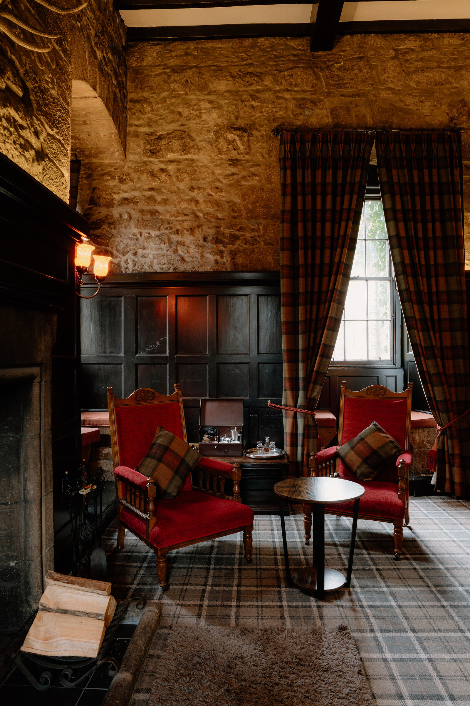 Red plaid armchairs and tartan curtains furnish a stone-walled sitting room beside a fireplace.
