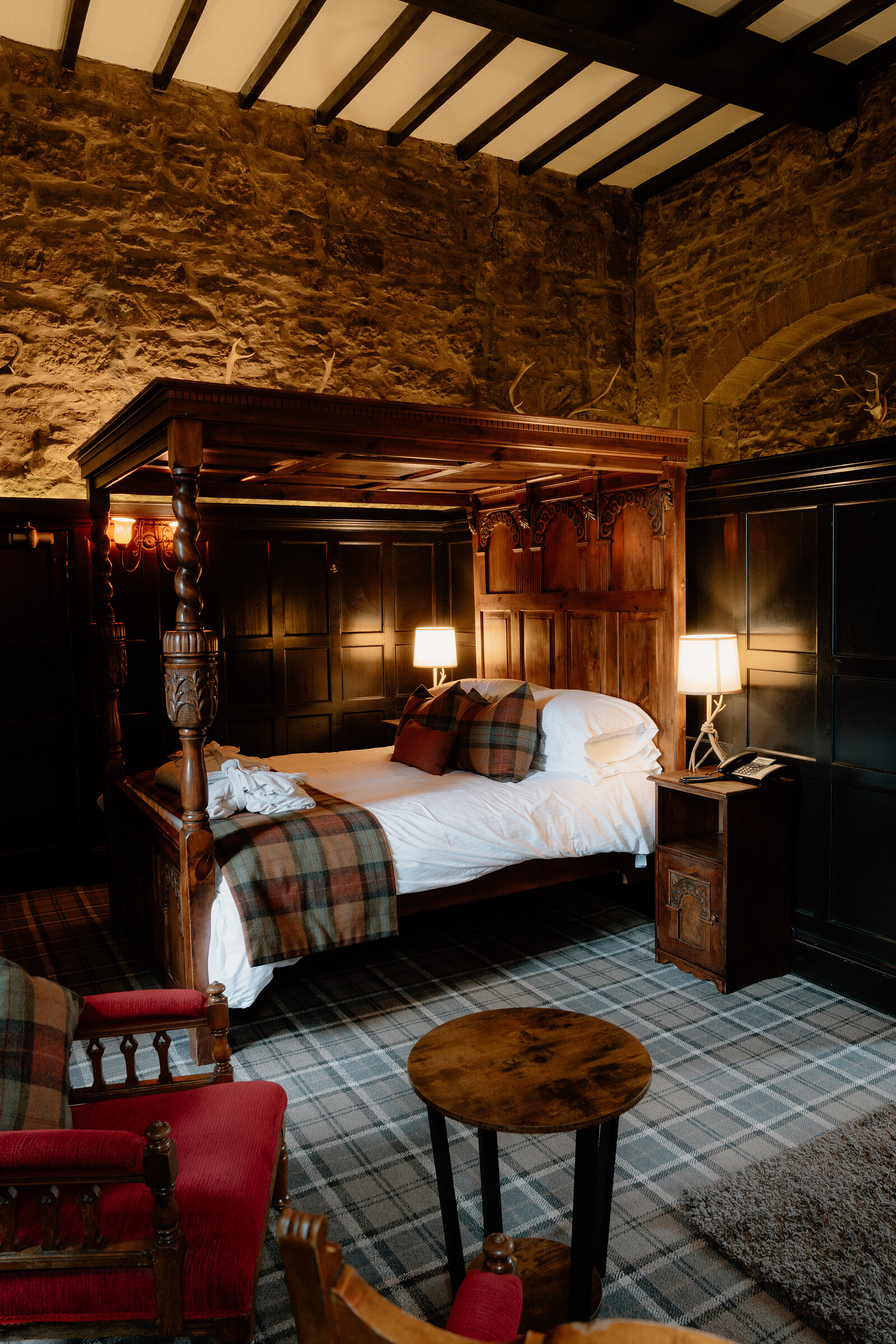 A carved four-poster bed with tartan throws stands against a stone wall beside red armchairs.