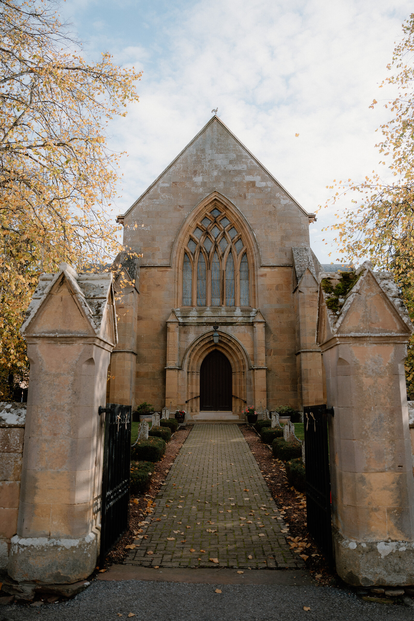A Gothic sandstone church stands beyond a gated path lined with autumn trees.