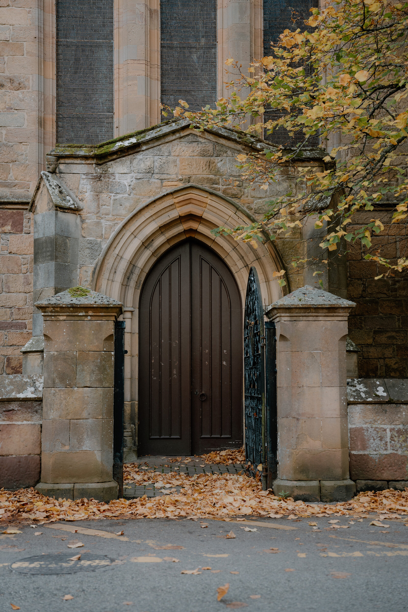 An arched wooden door sits beneath a Gothic church's stone gable, autumn leaves at its threshold.