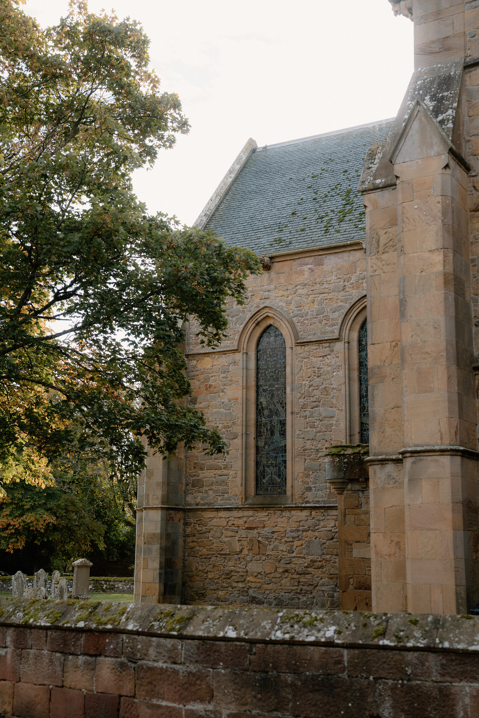 A Gothic sandstone church's buttressed wall and stained-glass window rise above a row of gravestones.