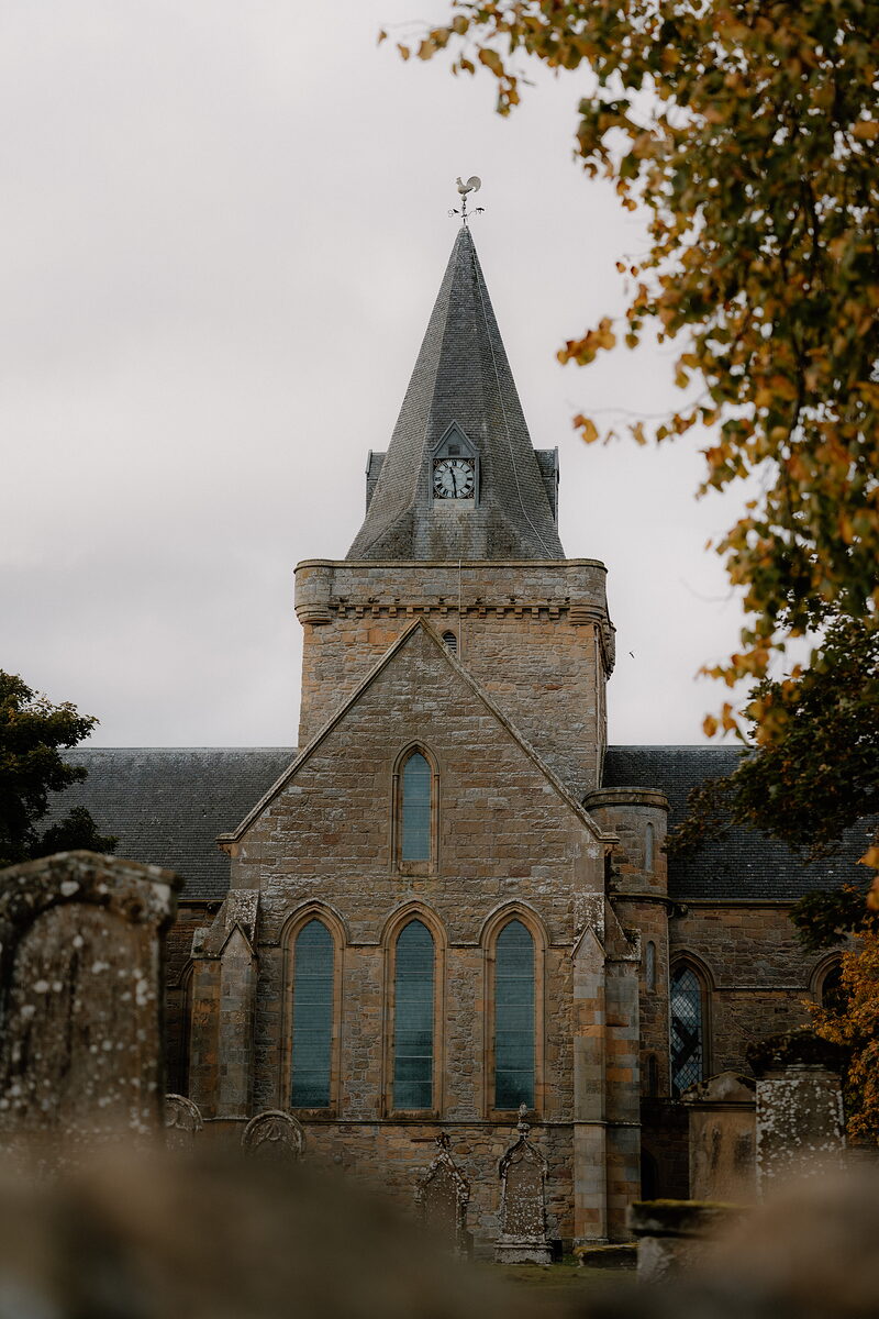 A church's spire and clock tower rise above weathered gravestones and an autumn tree.