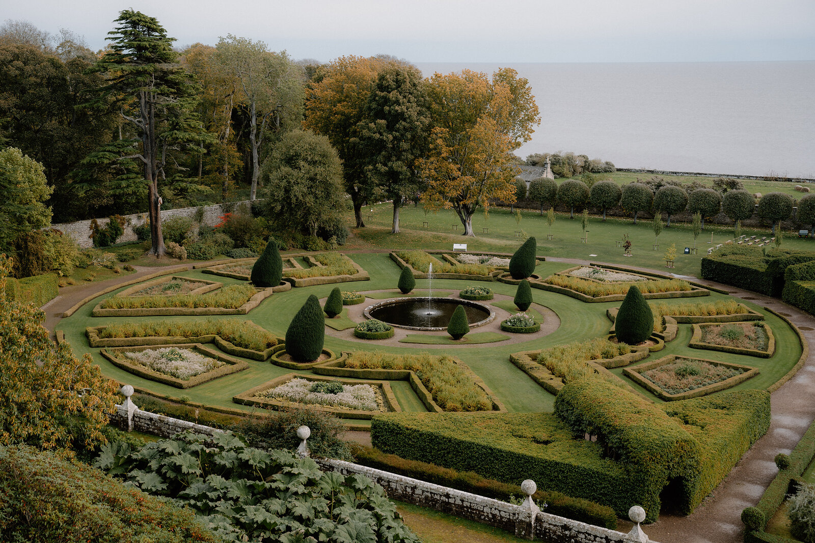 A circular fountain anchors a formal garden's geometric hedges beneath autumn trees by the sea.