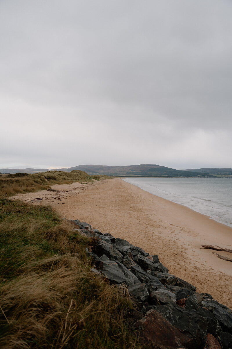 A wide sandy beach backed by dune grass, with hills rising along the far shore.