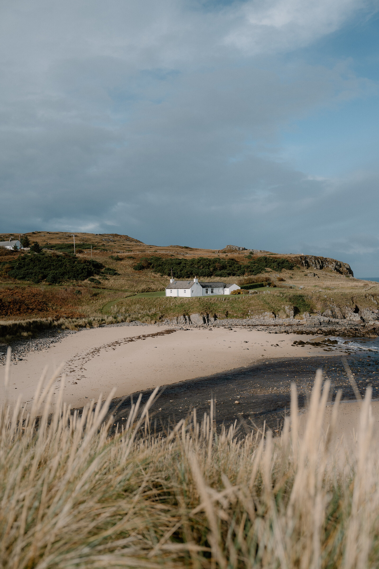 A white cottage set above a curved sandy bay, dune grass in the foreground