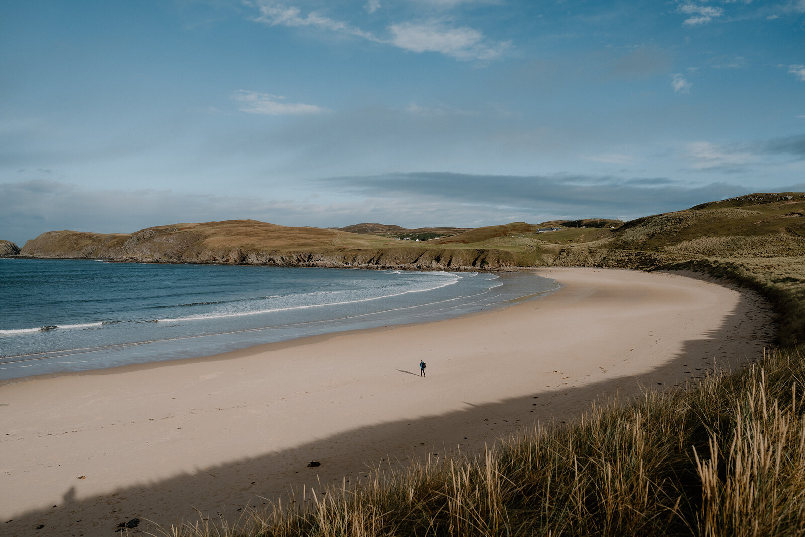 A lone walker crossing a wide curved beach backed by grassy dunes